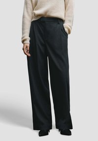 Pantalon large noir à fines rayures avec poches avant, fabriqué dans un tissu texturé, associé à un pull léger en maille.