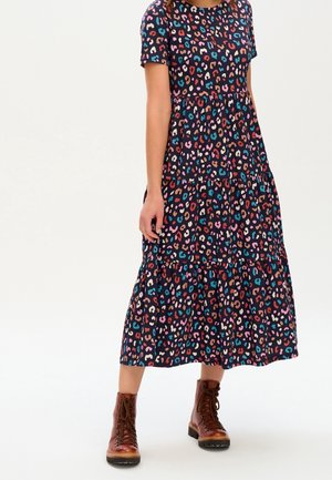 Vrouw draagt een zwarte, meerkleurige midi-jurk met luipaardprint, korte mouwen en bruine veterlaarsjes tot de enkel.