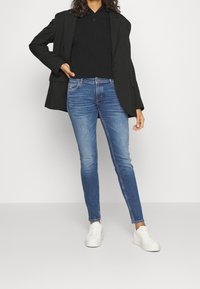 Person som bär svart blazer, svart skjorta, blå skinny jeans och vita sneakers, står med en hand på höften.