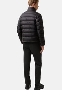 Mand iført sort pufferjakke, sorte bukser og sorte sneakers, som står med ryggen til mod en ensfarvet lys baggrund.