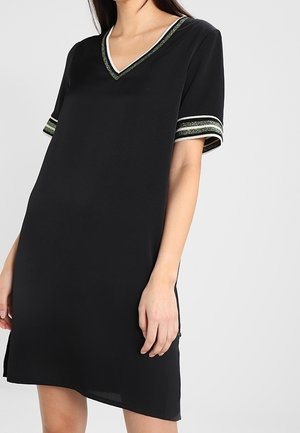 Femme portant une robe noire à manches courtes avec un décolleté en V et une bordure rayée brillante verte et blanche sur les manches et le col.