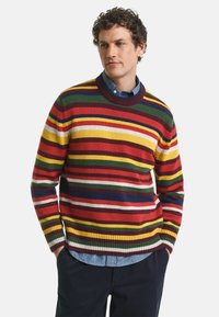 Multikleurige gestreepte trui met rode, gele, groene en marineblauwe patronen, ronde halslijn, geribbelde manchetten en zoom, gelaagd over een blouse met kraag.