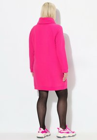 Vestido de punto fucsia con escote tipo cowl, mangas largas y puños acanalados; llevado sobre medias negras y zapatillas con acentos rosas.