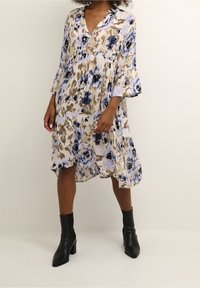 Robe fleurie en beige, bleu et noir avec décolleté en V et manches évasées. Dispose d'un ourlet à volants et d'un tissu léger. Bottines noires à la cheville.