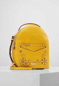 Mochila de cuero amarilla con adornos florales, forma redondeada, bolsillo frontal con cremallera y correa de hombro desmontable. Presenta un detalle del logo.