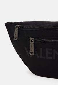 Svart nylontygs väska med en rundad form, utrustad med två metallblixtlås med logotypprydda dragband och en diskret logotyp tryckt på ytan.