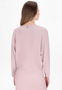 Maglietta a maniche lunghe rosa chiaro realizzata in materiale morbido e texturizzato. Presenta una vestibilità comoda e un collo rotondo, cuciture visibili e una piccola etichetta sul retro.