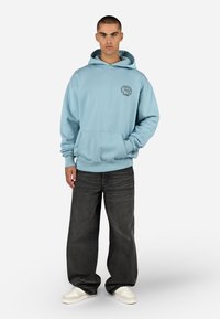 Ανοιχτό μπλε φούτερ με τσέπη καγκουρό, ribbed μανίκια και στρογγυλό λογότυπο στο στήθος. Φοριέται με χαλαρές σκούρες γκρίζες τζιν παντελόνες και λευκά αθλητικά παπούτσια.