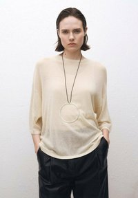 Beżowy, półprzezroczysty, oversize sweter z rękawami do trzech czwartych, zestawiony z czarnymi, plisowanymi spodniami i dużym, srebrnym naszyjnikiem w kształcie okręgu.