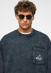 Donkergrijze sweatshirt met een textuurfinish, voorzien van een patchzak met "Atelier" geborduurd in het wit. Model draagt zwarte zonnebril.