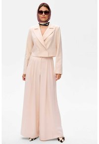 Lichtroze, dubbelzijdig gefacetteerde, cropped blazer met bijpassende wijde broek. Model draagt donkere zonnebril en een gestructureerde sjaal. Gladde textuur.