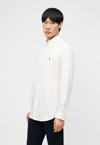 Muž oblečený v bielej košeli s dlhými rukávmi a čiernych nohaviciach, stojaci na jednoduchom svetlom pozadí.