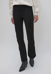Pantalon noir évasé avec une texture lisse et une taille élastique, associé à des bottines noires, mettant en valeur une silhouette élégante.