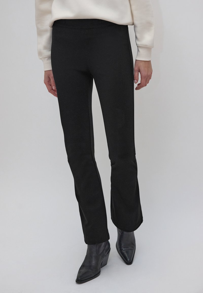 Pantalon noir évasé avec une texture lisse et une taille élastique, associé à des bottines noires, mettant en valeur une silhouette élégante.