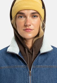 Ungdom med blå øyne som har på seg en gul strikket lue, brun hettegenser og blå denimjakke med hvit fleecekrage, ser rett frem.