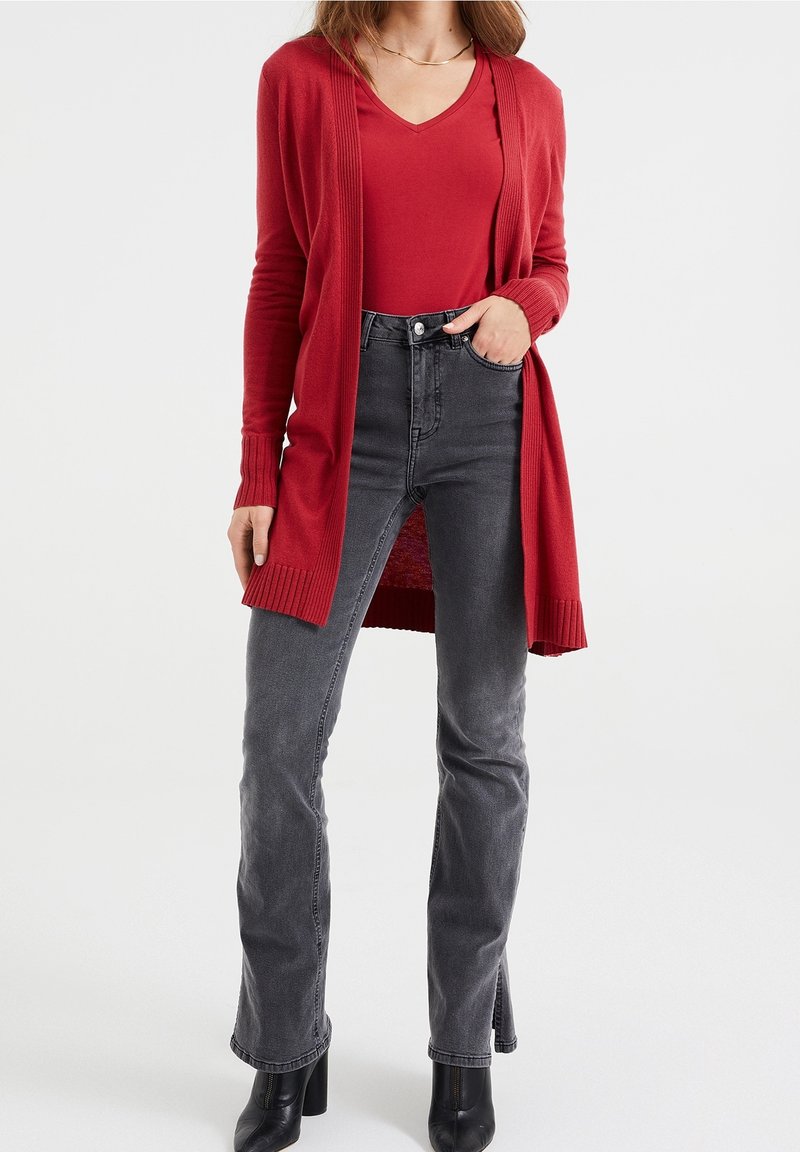 Long cardigans rouges avec des poignets côtelés, porté sur un top rouge à col en V, associé à un jean gris foncé taille haute et des bottines à talons noires.