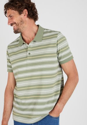 Uomo sorridente, indossa una polo a righe verdi a maniche corte e jeans blu, con una mano in tasca, guarda verso sinistra.