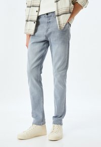 Jeans de mezclilla azul claro con un corte ajustado, que presentan un desgastado sutil y un diseño de cinco bolsillos, combinados con zapatillas de deporte beige.