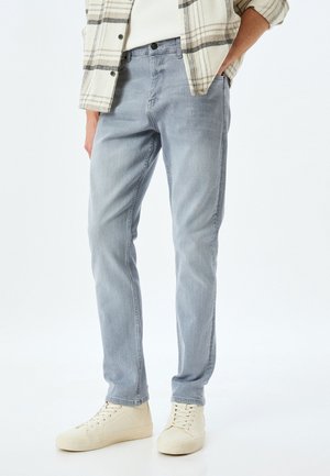 Lyseblå denimjeans med slim fit, der har diskrete slidmærker og fem-lomme design, båret med beige sneakers.