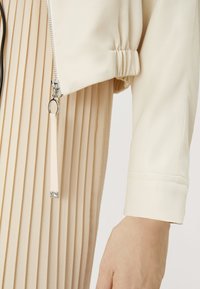 Giacca beige con zip, finitura testurizzata e dettagli plissé. Presenta una zip argentata e un laccetto in tessuto attaccato all'orlo.