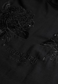 Tela negra con patrones florales bordados y lentejuelas. Texto "superdry losangeles" en hilo negro debajo del diseño.