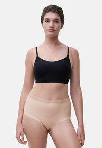 Frau steht in einem schwarzen, kurz geschnittenen Camisole und beigen Unterwäsche mit hohem Bund und Spitzenbesatz vor einem schlichten hellen Hintergrund.