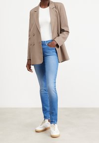Blazer beige de doble botonadura sobre una camiseta blanca ajustada, combinado con vaqueros azul claro y zapatillas blancas con suela marrón claro.