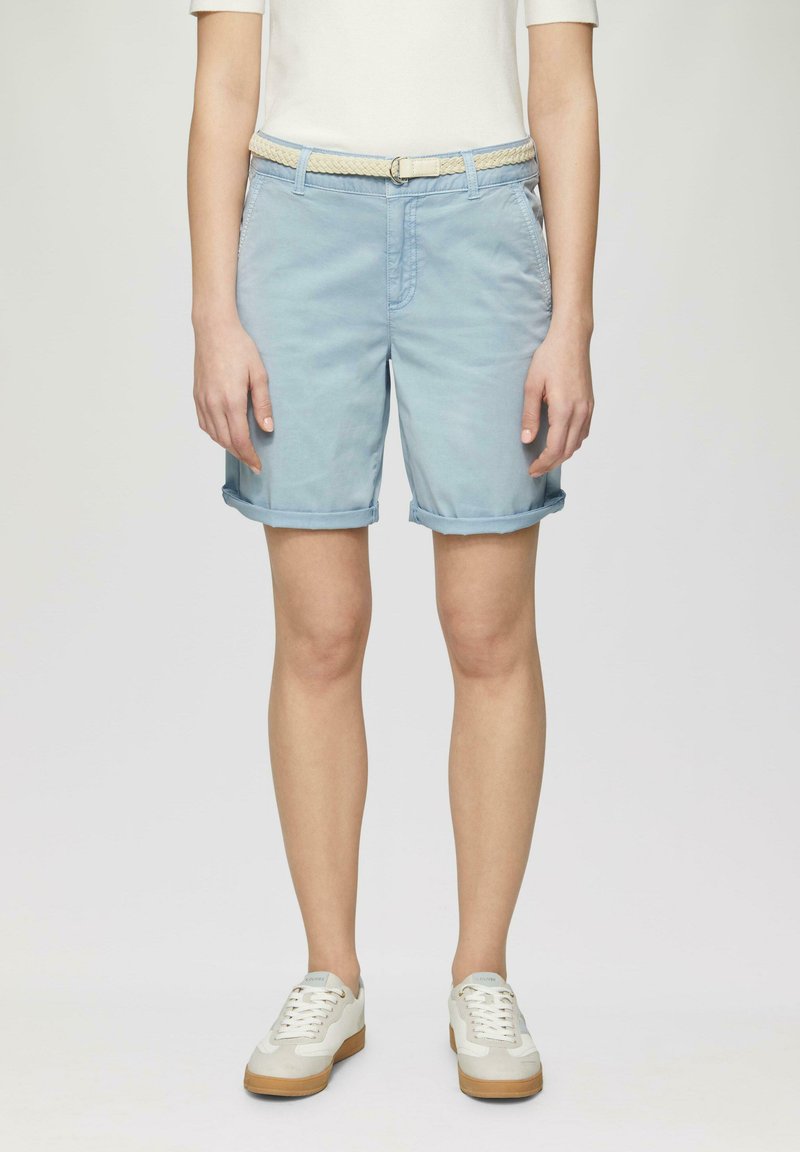 Shorts bleu clair en coton avec des ourlets retournés, dotés de poches latérales et d'une ceinture tressée beige. Portés avec des sneakers blanches et beiges.