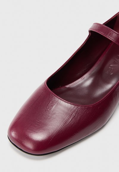 Scarpa Mary Jane in pelle bordeaux con punta quadrata, texture liscia e una sottile cinghia sopra il collo del piede. La suola presenta una leggera curva.