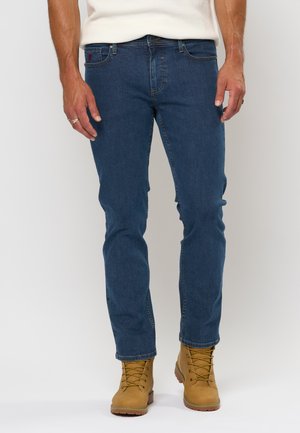Jean en denim bleu avec une coupe slim, comportant cinq poches et des détails en couture. Porté avec des bottes de travail marron, mettant en valeur une texture robuste.