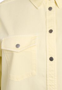 Svetložltá rifľová košeľa s vreckom na hrudi a chlopňou zabezpečenou kovovým patentným gombíkom, s prednou légou so tromi viditeľnými patentnými gombíkmi.