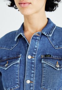 Chemise en denim bleu moyen avec un col pointu, des boutons argentés et deux poches poitrine. Coutures orange notables et détail de patch logo.