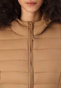 Beige, gepolsterte Weste mit einem Reißverschluss vorne, Kapuze und horizontaler Nähte. Das Material erscheint glatt mit einem matten Finish.
