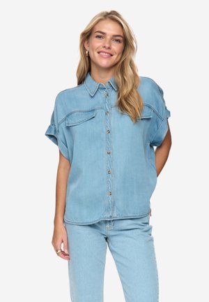 Lächelnde Frau mit langen blonden Haaren trägt ein hellblaues Denimhemd mit kurzen Ärmeln und passende Jeans, steht vor einem schlichten Hintergrund.