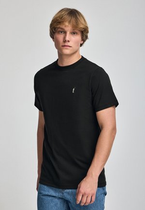 T-shirt nera in cotone con maniche corte, scollatura rotonda e ricamo del logo discreto. Abbinata a jeans in denim azzurro chiaro.