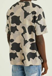 Camiseta de manga corta en beige con un patrón de camouflage abstracto en negro y gris, con cuello redondo y corte relajado.
