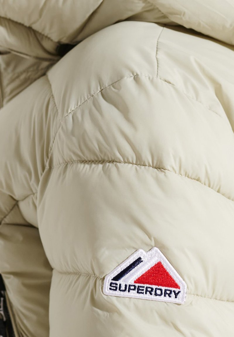 Sezione di giacca puffer beige con texture trapuntata e un'etichetta con il marchio. Presenta un design triangolare rosso, bianco e blu sulla manica.