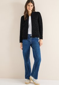 Zwarte bomberjack met een gestructureerd oppervlak, een ritssluiting aan de voorkant en een ronde kraag, gecombineerd met rechte blauwe jeans en beige sneakers met blauwe accenten.