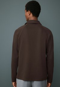 Camisa marrón de manga larga con cuello, de tela texturizada y mangas raglán. Diseño sencillo, dobladillo recto, con costuras en contraste.