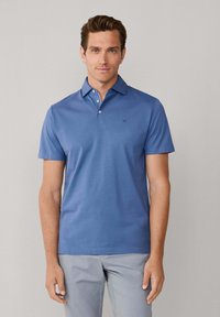 Polo bleu avec une texture douce, manches courtes, col et trois boutons. Présente un petit logo sur la poitrine. Porté avec un pantalon gris.