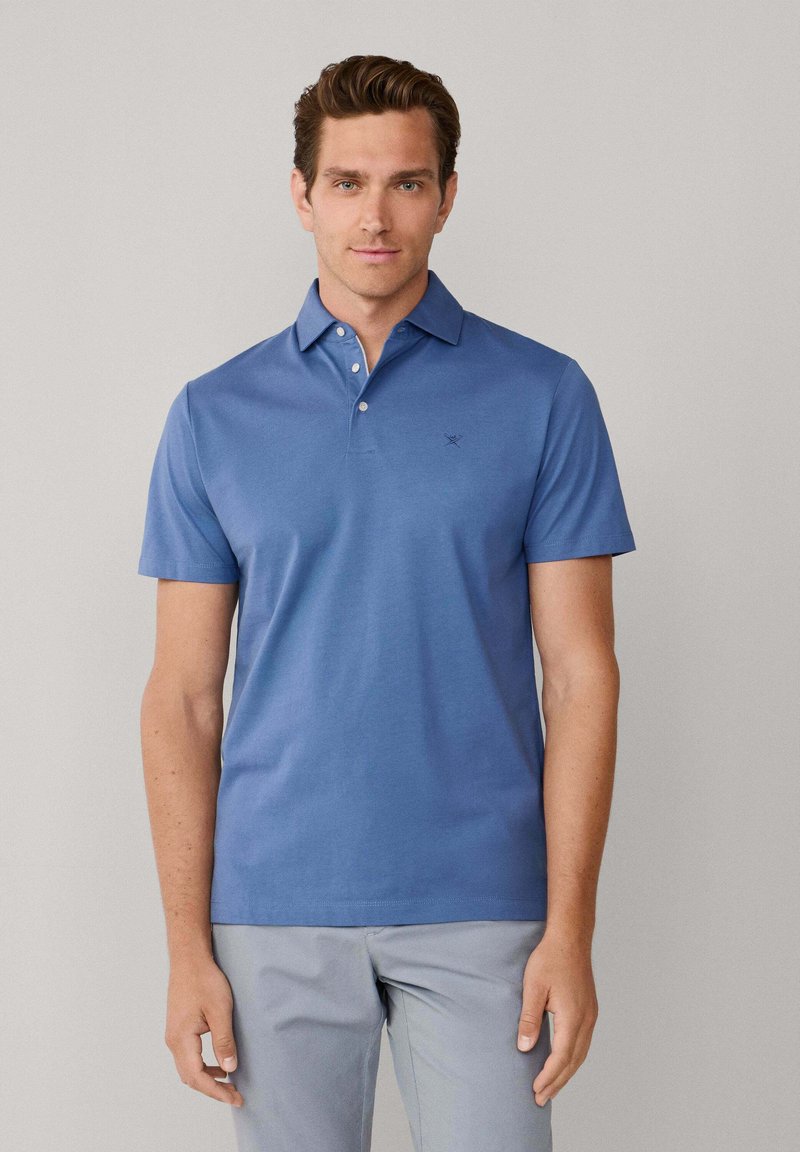 Polo bleu avec une texture douce, manches courtes, col et trois boutons. Présente un petit logo sur la poitrine. Porté avec un pantalon gris.