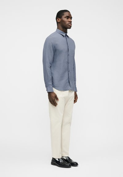 Άνδρας όρθιος, φοράει ανοιχτό μπλε πουκάμισο με κουμπιά, κρεμ παντελόνι και μαύρα επίσημα παπούτσια, με φόντο απλό λευκό.