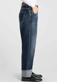 Jean en denim bleu foncé avec une coupe droite, présentant des ourlets retroussés, un design à cinq poches et des accents en métal doré.