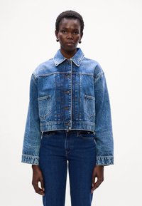 Giacca di jeans corta in azzurro chiaro, con chiusura a bottoni, grandi tasche e colletto classico. Texture con cuciture visibili e effetti di sbiadimento.