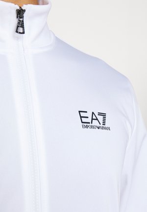 Primo piano di una giacca bianca con zip e colletto alto con logo nero EA7 Emporio Armani sul petto.