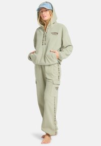 Hellgrüner Fleece-Overall mit Kapuze, Reißverschluss, Taschen und gemusterten Akzenten an den Seiten. Dazu eine blaue Mütze.