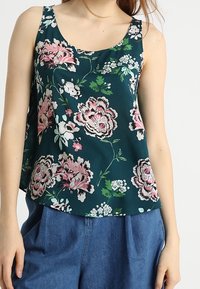 Débardeur floral en bleu canard foncé avec des fleurs rose et blanche. Tissu léger, décolleté en forme de scoop et coupe ample. Associé à un pantalon taille haute bleu.