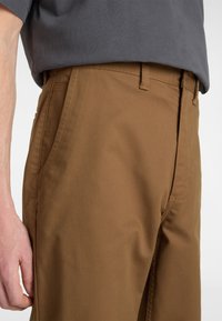 Braune Baumwollhosen mit einem geraden Bein Design, ausgestattet mit Seitentaschen und Gürtelschlaufen. Die glatte Textur und minimale Nähte sind sichtbar.