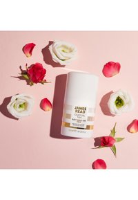 Bottiglia cilindrica bianca di James Read Gradual Tan, 50ml, con accenti dorati e testo nero, circondata da petali di rose rosa e bianchi su uno sfondo rosa.