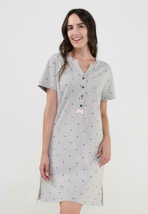 Mujer joven sonriendo, vestida con un camisón gris de manga corta con pequeños corazones negros, botones en la parte delantera y un pequeño lazo rosa cerca de la cintura.